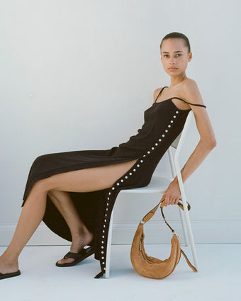 Image of model sitting in white metal chair wearing Astrid Knit Dress in Boucle Viscose in black, carrying Stanton Sling in Suede in honey