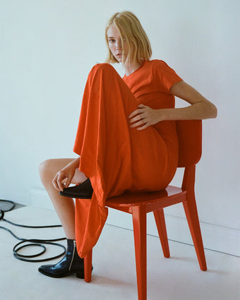 Image of model sitting on red chair, wearing Noelle Dress in Jersey in poppy